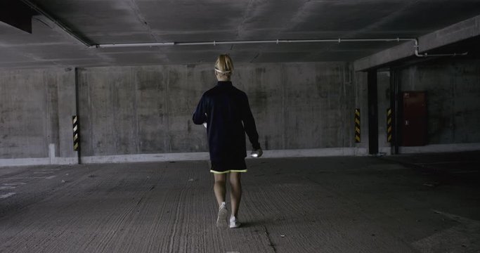 TRACKING Teenager girl soccer player walking towards a wall in empty covered parking garage, holding spray paint can in her hands. 4K UHD 60 FPS RAW graded footage