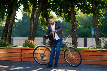 Obraz premium Bearded Businessman in business suit riding on retro bicycle to work on urban street in the morning on sunset. Drink coffee to go and speaking on mobile phone