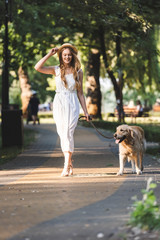 full length view of beautiful girl in white dress and straw hat smiling while walking with golden retriever on pathway and looking away