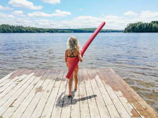 Caucasian preschool blonde girl in pink swimsuit standing on wooden lake river dock with pool noodle on summer day. Happy healthy active lifestyle childhood. View from back behind.