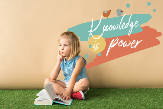 Thoughtful Cute Child Sitting With Crossed Legs On Grass Rug And Holding Book On Beige Background With Knowledge Is Power Lettering