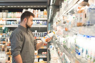 Man is in the milk department of a supermarket, holding a bottle of milk in his hand and reading a...