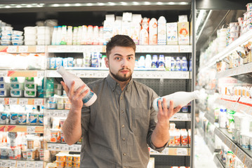 Man is standing with two bottles of milk in the milk department of the supermarket and can not...