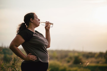 Overweight woman in sportswear drinking water after outdoor workout. Healthy lifestyle, hydration, weight loss, activity concept