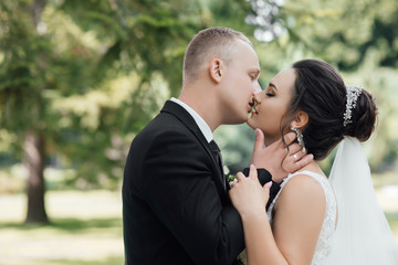 Stylish couple of happy newlyweds posing in the park on their wedding day. Perfect couple bride, groom posing and kissing. Young wedding couple enjoying romantic moments.