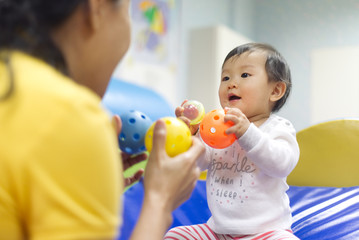 Young little Asian baby holding balls and toys playing with female preschool teacher in classroom....