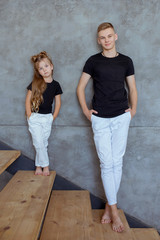 stylish caucasian siblings (brother and sister) in blak t-shirts and white pents standing by the gray wall in loft interior. Family, fashion, relatives, relationship and children concept.