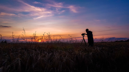 Silhouette of the photographer with digital camera and a tripod taking pictures of the sunset in the field