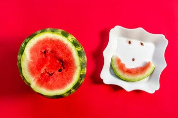 A cheerful smiling watermelon, a face of slice and pips on a white plate and a large ripe on a red background. The view from the top, flat lay. The concept of a fun hot summer and good mood