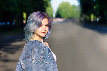 Beautiful girl teenager with multicolor dyed purple and blue hair in jeans jacket standing outdoor in summer park at a sunny summer day, looking at camera. Young stylish woman walking outside, evening