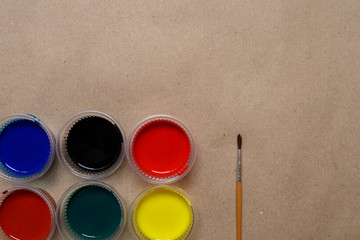 Many water colors are available in cups. Colors are red, blue, green, yellow, brown, black and brush on the background of brown paper.