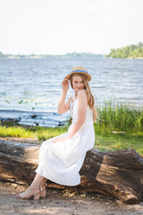 Fototapeta na wymiar beautiful girl in white dress and straw hat sitting on trunk of tree on river shore and looking away
