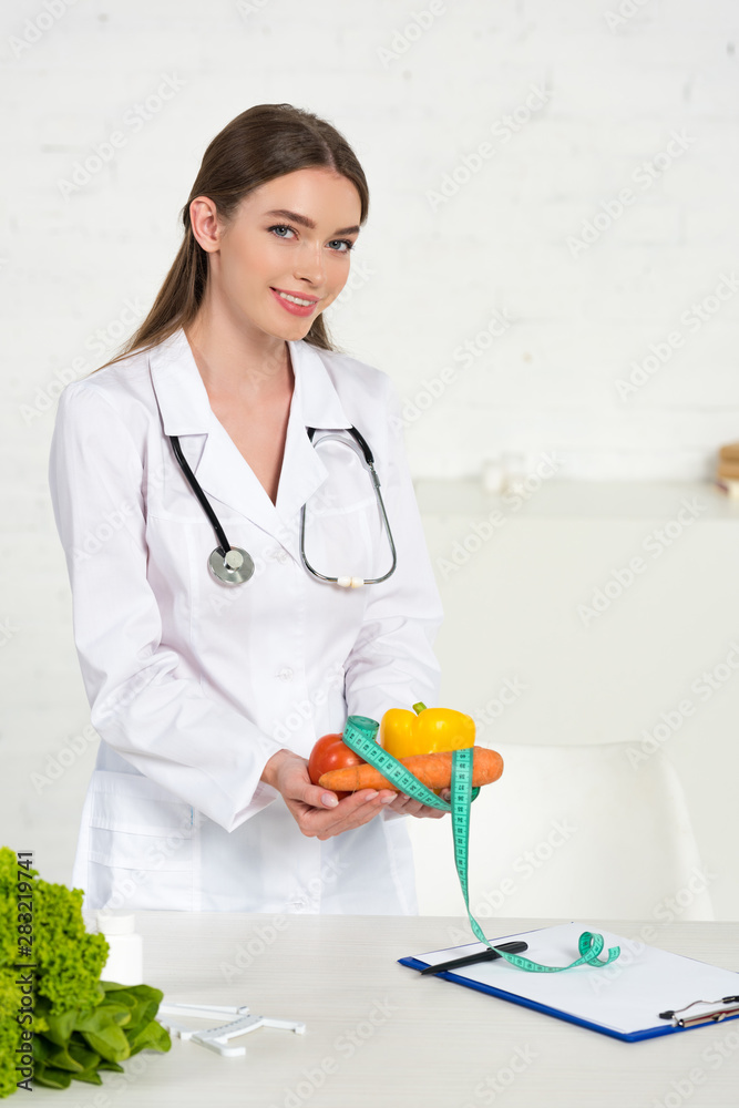 Wall mural dietitian in white coat holding vegetables and measure tape at workplace