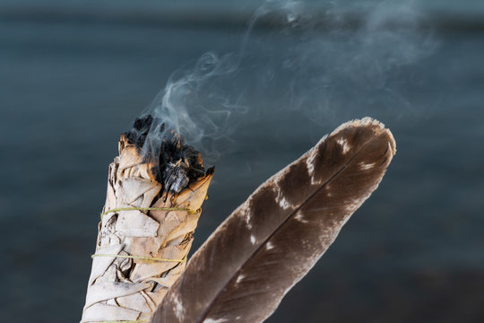 Smudging Ritual using burning thick leafy bundle of White Sage Grade A barred Turkey Smudging Feather on the beach at sunrise in front of the lake.