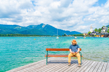 Middle aged male sitting on a bench on wooden deck by the Lake St. Wolfgang, Austria. St. Walfgang market town in the background.