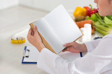 cropped view of dietitian in white coat reading book at workplace