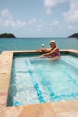 Smiling middle-aged woman in swimsuit with cocktail enjoys swimming pool by the turquoise sea