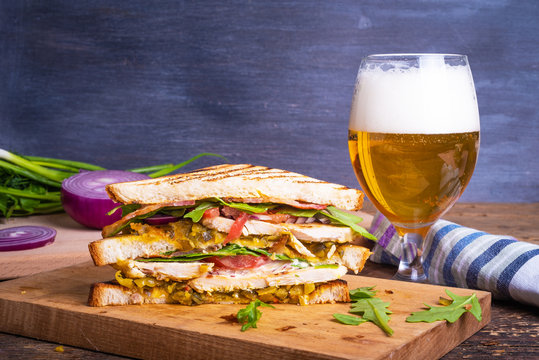 Tasty triangular sandwich made from toasted bread with chicken breast, fried bacon, vegetables, pickles with mustard and arugula closeup and a glass of beer with foam on rustic background, copy space