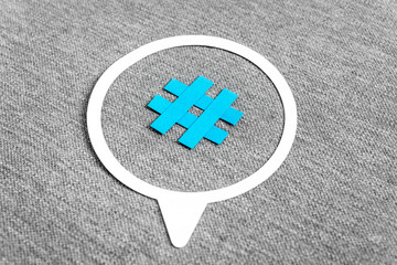 Paper cutout of blue hashtag symbol on a speech bubble on grey textured background. Concept of social media and digital marketing.