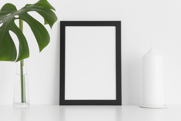 Black frame mockup with a candle and a monstera leaf in a vase on a white table.Portrait orientation.