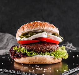 Tasty burger with mozzarella cheese, tomatoes, lettuce and sauce pesto on slate black background,...