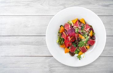 Healthy salad with seared tuna fish, greens, bell pepper and sauce in plate on light wooden background. Healthy food, clean eating, dieting, top view