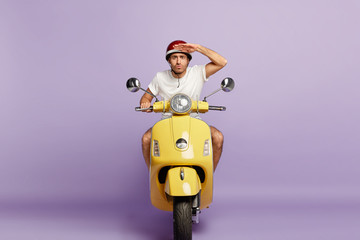 Serious motorbike driver keeps palm near forehead, concentrated into distance, being attentive on road, wears protective headgear, being excellent motorcyclist, isolated on purple background