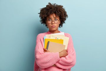 Shocked Afro American university student stands with textbooks, afraids of passing exam, dressed...