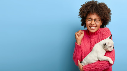 Joyful dark skinned woman poses with cute black and white puppy, rejoices its recovering after...