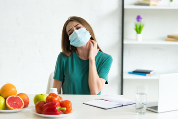 young woman in medical mask sitting at table with fruits, vegetables and pills and scratching neck at home