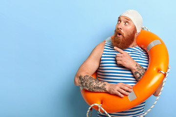 Surprised unshaven male vacationist dressed in bathingcap and striped vest, poses with swimring, points aside with index finger and looks surprisingly aside on blank space for your promotion