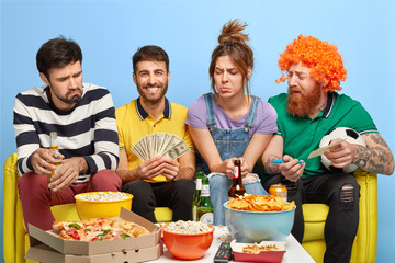 Company of friends watch soccer on TV, one guy makes bet for winning his favourite team, holds money, sit at couch, eat junk food and drink beer. Friendship, sport and entertainment concept.
