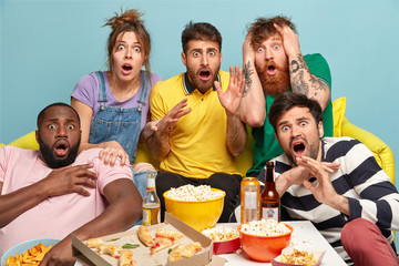 Scared group of friends watch horror movie, feel great fear and surprise during thrilled scary film moment, have bated breath, eat junk food, spend lazy day off together sit on couch against blue wall