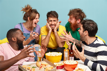 Frustrated guy plugs ears, avoide noise, doenst want to hear friends who shout loudly with irritation at him, spend free time at home watching movie distracted from TV set during television advertsing