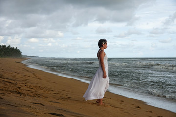 Fototapeta na wymiar Girl in a white dress meets the sunset on the beach of the tropical ocean. Young girl looks into the distance
