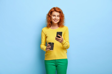 Pretty ginger female holds modern mobile phone and types message, drinks takeout coffee, enjoys leisure time, searches needed information in internet, dressed in vibrant outfit. Online chatting