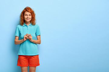 Lovely smiling female keeps hands together and looks with intrigue, has toothy smile, wears comfortable summer clothes, waits for something pleasant, stands over blue wall with copy space on right