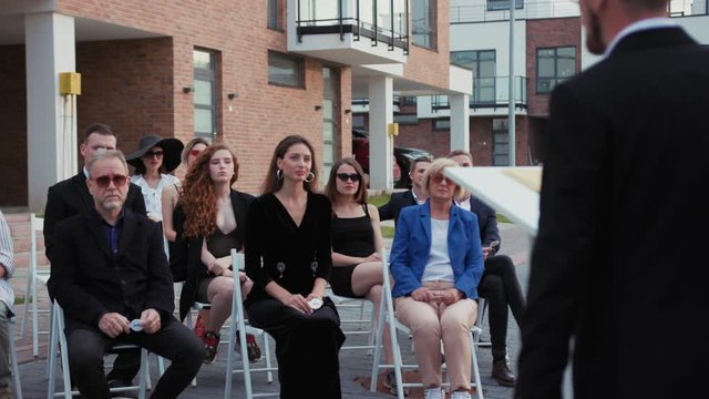 Appealing multi-age weaathy people seating in seats outdoors listening to the auctioneer. At modern property sale auction people bidding and buying real estate.