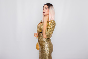 Charming young blonde haired woman wearing elegant gold evening dress and holding gold color clutch...