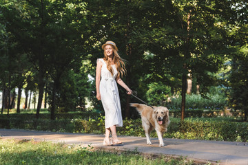 full length view of beautiful girl in white dress and straw hat walking with golden retriever