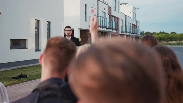 Fun auction sale. Back view of rich bidders raising hands bidding for real estate objetcs sitting outdoors. Charismatic male auctioneer leading a property auction.