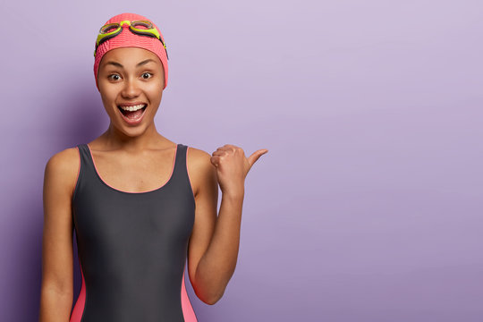 Joyful dark skinned female swimmer wears bathingcap, black swimsuit, smiles broadly, shows direction to swimming pool, spends leisure time at swim center, points thumb on copy space over purple wall
