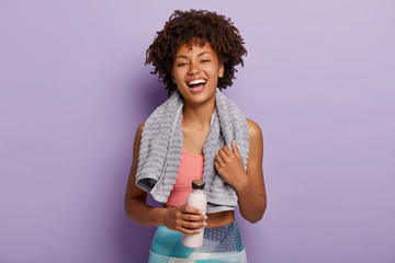 Smiling fitness woman in top and leggings takes break after training, holds bottle of water, wipes...