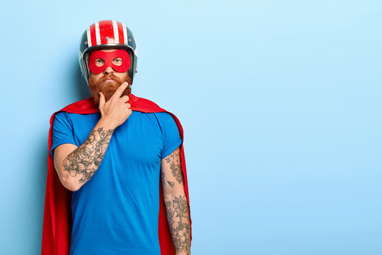 People and super power concept. Serious man with red thick beard, wears helmet and red superhero cape, has tattooed arm, looks confidently at camera, stands against blue wall, copy space area on right