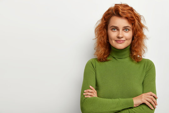 Studio shot of attractive young female model with curly short ginger hair, healthy skin, has gentle smile, dimples on cheeks, listens attentively something, keeps arms folded, wears green jumper