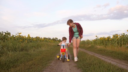 Mom teaches daughter to ride a bike on country road in a field of sunflowers. a small child learns to ride a bike. Mother plays with her little daughter. The concept of happy childhood.