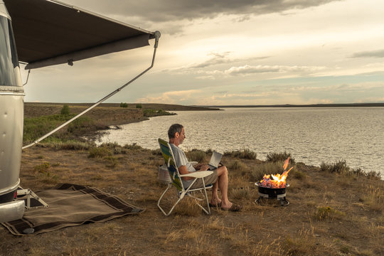 A programmer out of office, working remotely at a camping spot by a lake, campfire burning and a camper near by