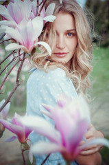 Beautiful happy young woman enjoying smell in a flowering spring garden.Beutiful tree magnolias,big flowers. Blonde with blue eyes and blue dress.