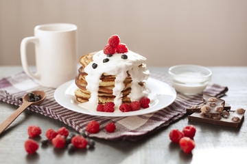 Stack of pancackes with sour cream and fresh berries, mug of tea or tea on dark surface, side view of table decorated with muffins, raspberries, blueberries, chocolate. Breakfast on cotton towel.