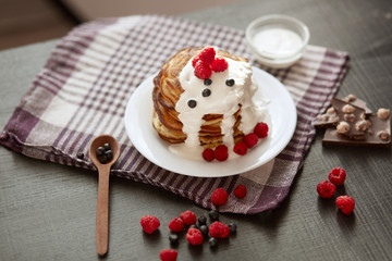 Picture of stack of pancakes with blueberries and raspberries on white plate, dark brown colour r backdrop decorated with cup of tea, spoon, pieces of chocholate and berries. Delicious breakfast.